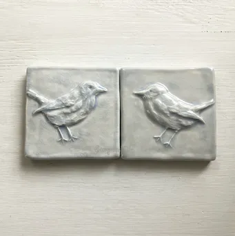 Two ceramic tiles featuring intricately embossed birds, showcasing a soft gray finish. Ideal for decorative purposes or wall art.