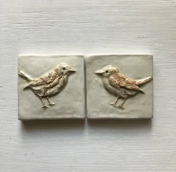 Two ceramic tiles featuring bas-relief designs of birds, showcasing detailed textures and subtle color variations. Ideal for home decor or artistic display.