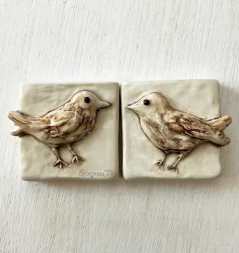 Two ceramic tiles featuring reliefs of birds, showcasing intricate details and a soft color palette, perfect for decorative accents.