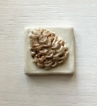 Ceramic tile with a textured, organic shape resembling a pine cone, showcasing earthy tones and intricate details, ideal for decorative purposes.