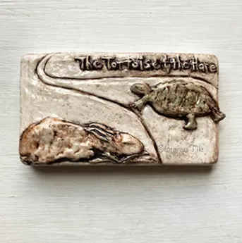Ceramic tile featuring a tortoise and a hare, illustrating the fable "The Tortoise and the Hare." The embossed design emphasizes the moral of perseverance.