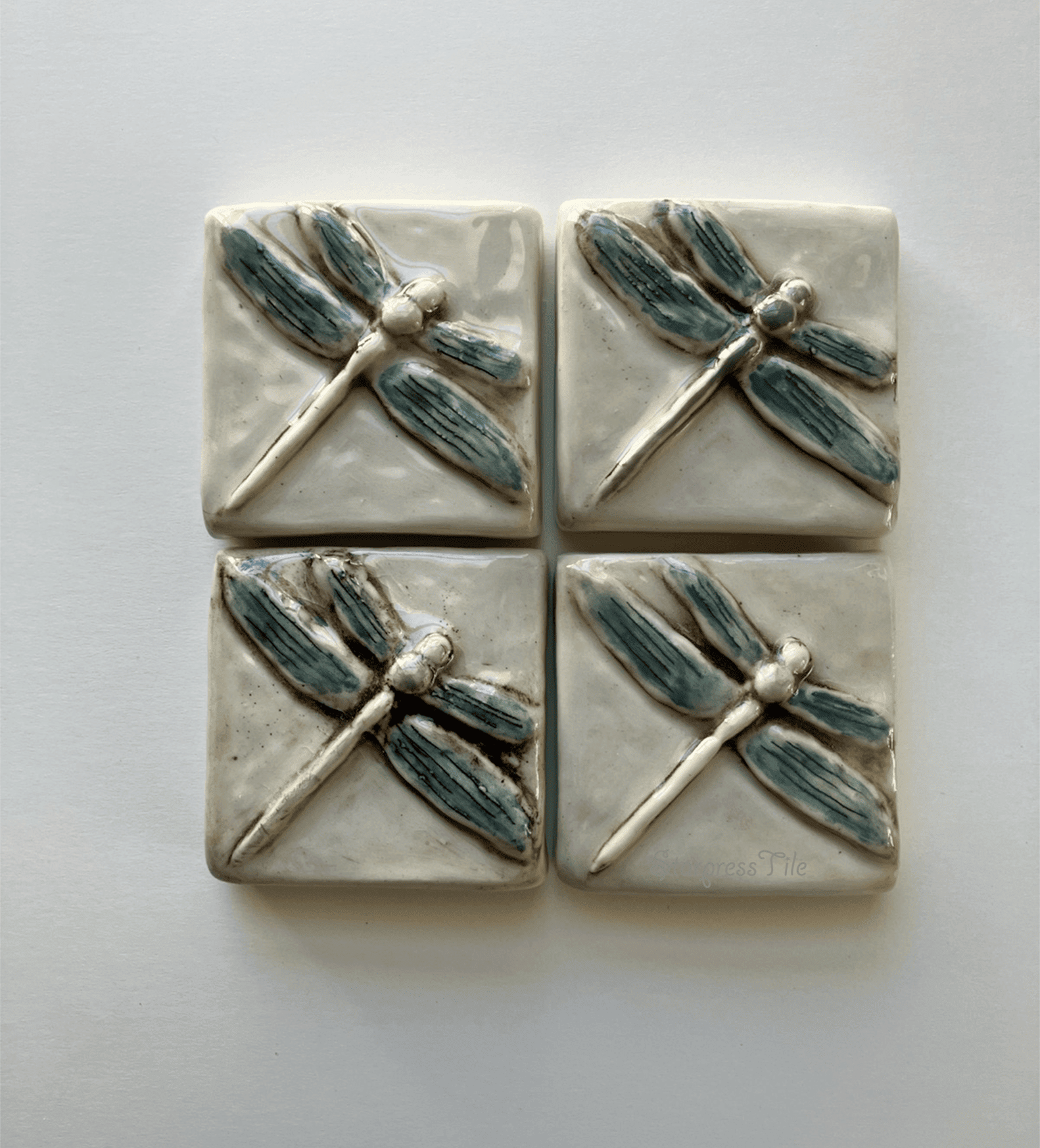 Four ceramic tiles featuring embossed dragonfly designs in a watercolor hand painted teal and brown highlight. The artwork adds a natural element to home decor.